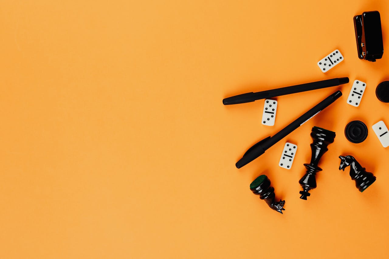 services-04 A minimalist arrangement of chess pieces, dominoes, pens, and a stapler on a vibrant orange background.